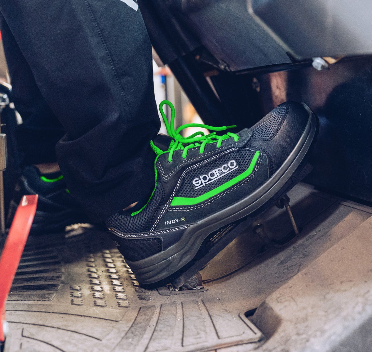 A close-up of a person's foot wearing a black Sparco racing shoe with bright green laces and trim, positioned over a motorcycle's foot pegs and brake pedal.