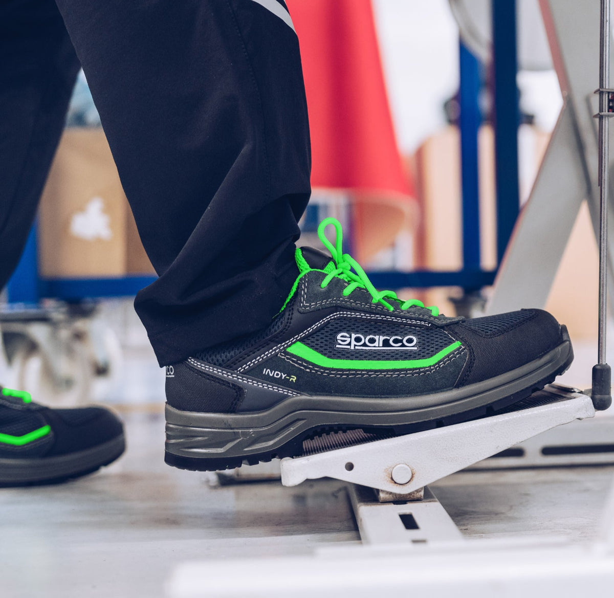 A close-up of a person's foot wearing a Sparco safety shoe while stepping onto a white metal platform. The shoe is black with bright neon green laces and green accents, paired with black trousers.