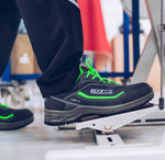 A close-up of a person's foot wearing a Sparco safety shoe while stepping onto a white metal platform. The shoe is black with bright neon green laces and green accents, paired with black trousers.