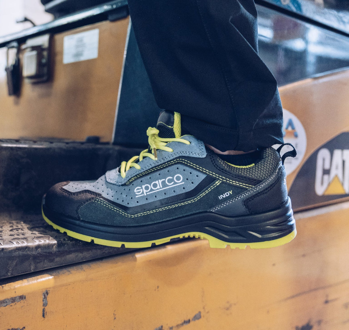 A close-up of a person's foot wearing a Sparco work shoe, standing on a yellow industrial machine. The shoe is dark gray and black with bright yellow laces and a yellow outsole.