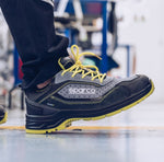 A Sparco safety shoe in dark gray and black with bright yellow laces and a yellow outsole, shown being worn with dark pants in an industrial setting.