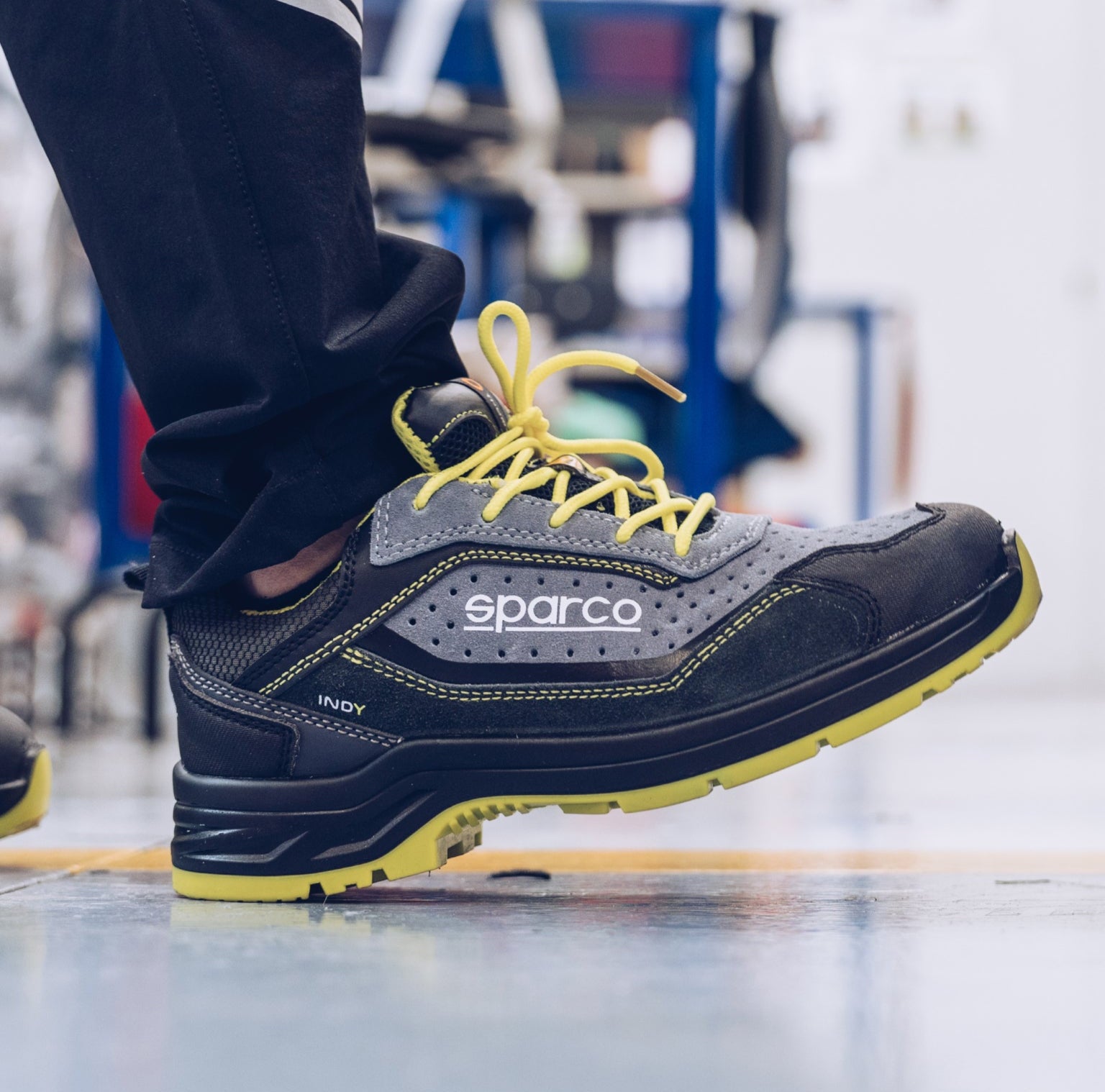 A Sparco safety shoe in dark gray and black with bright yellow laces and a yellow outsole, shown being worn with dark pants in an industrial setting.