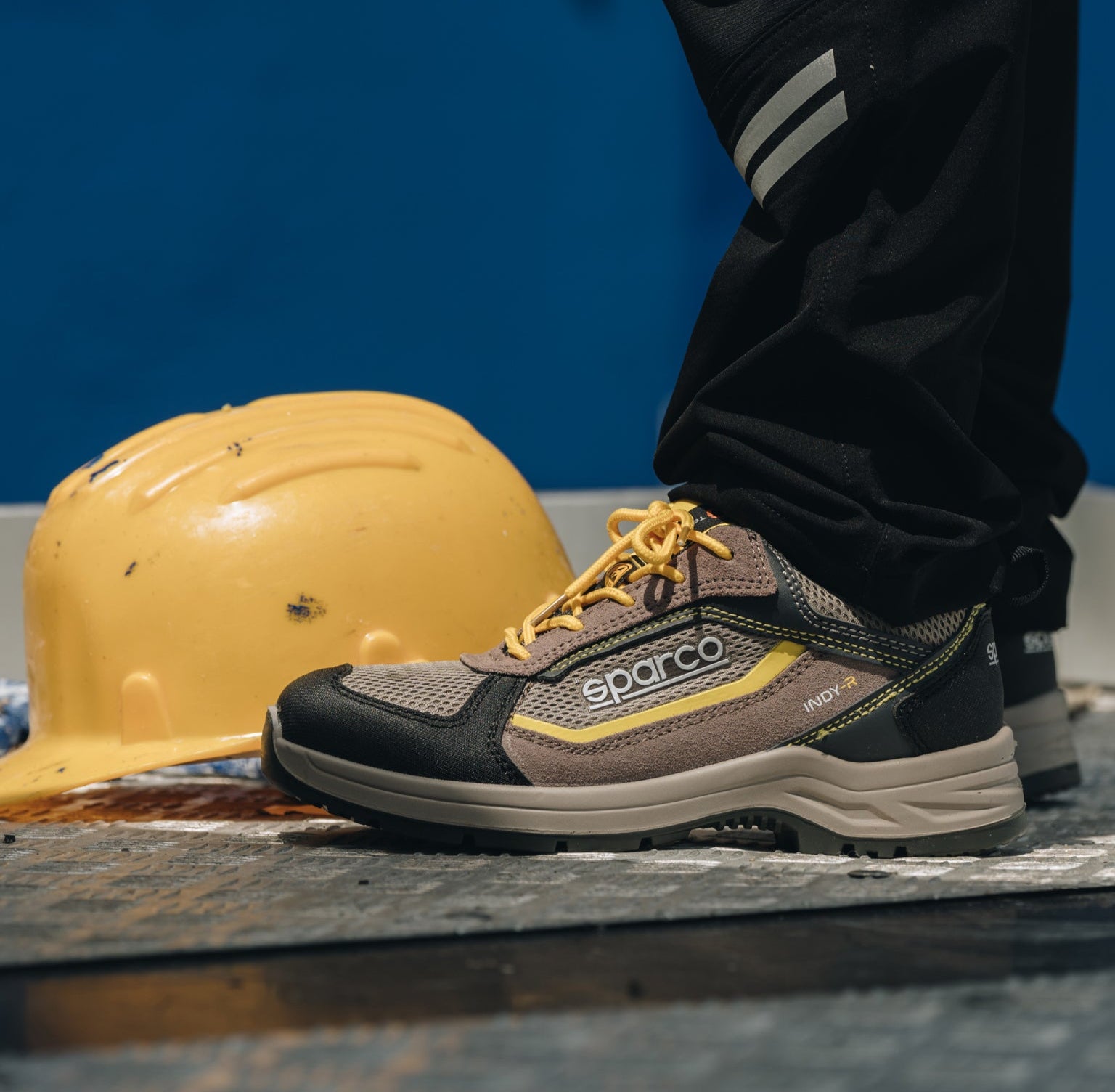 A Sparco work shoe in brown, gray, and yellow, shown on a person's foot next to a yellow hard hat against a blue background.