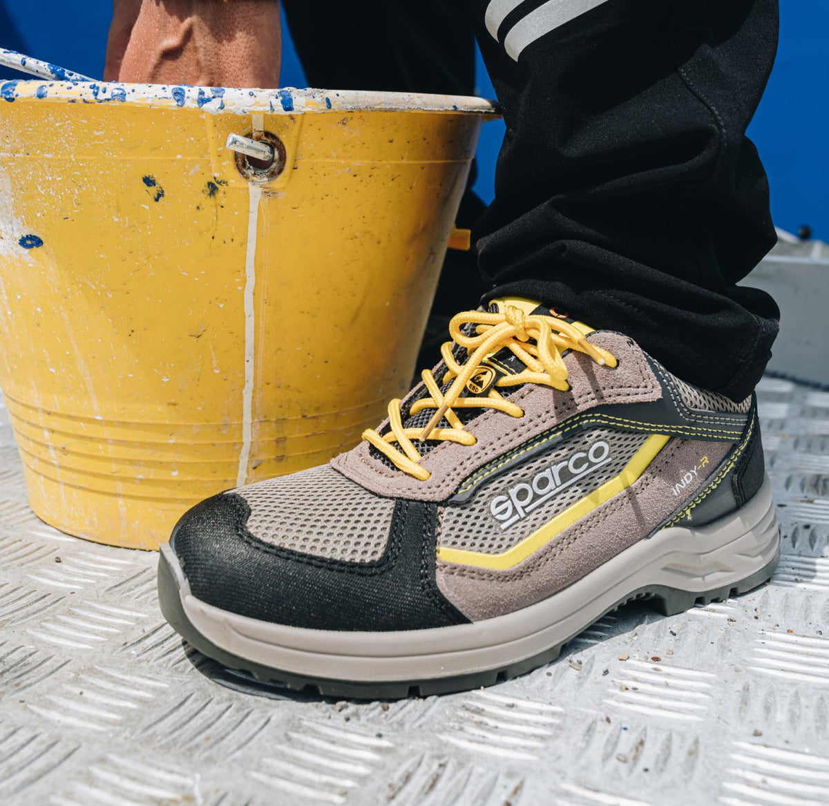A Sparco work shoe in gray and black with yellow laces, shown on a metal grate next to a yellow paint bucket.