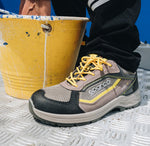 A Sparco work shoe in gray and black with yellow laces, shown on a metal grate next to a yellow paint bucket.