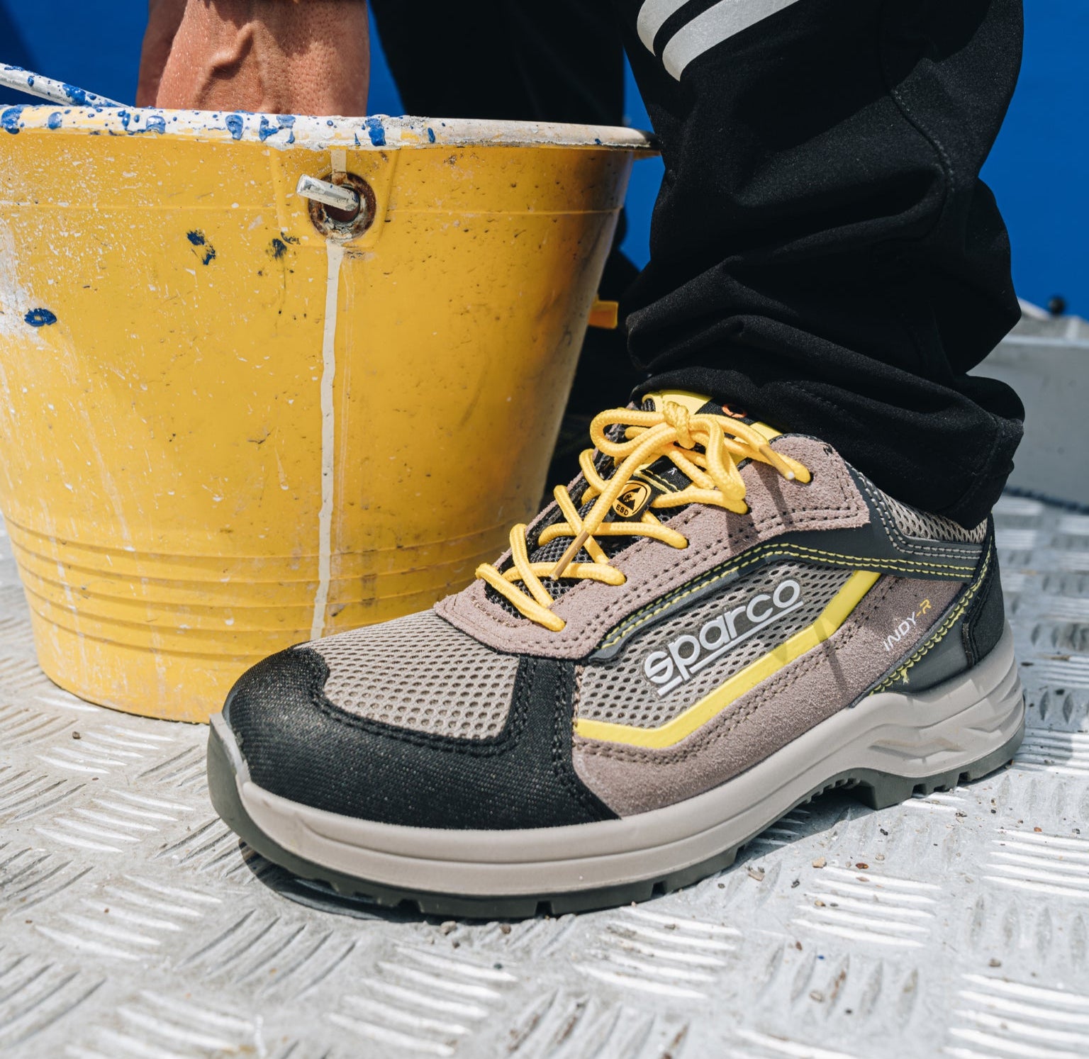 A Sparco work shoe in gray and black with yellow laces, shown on a metal grate next to a yellow paint bucket.