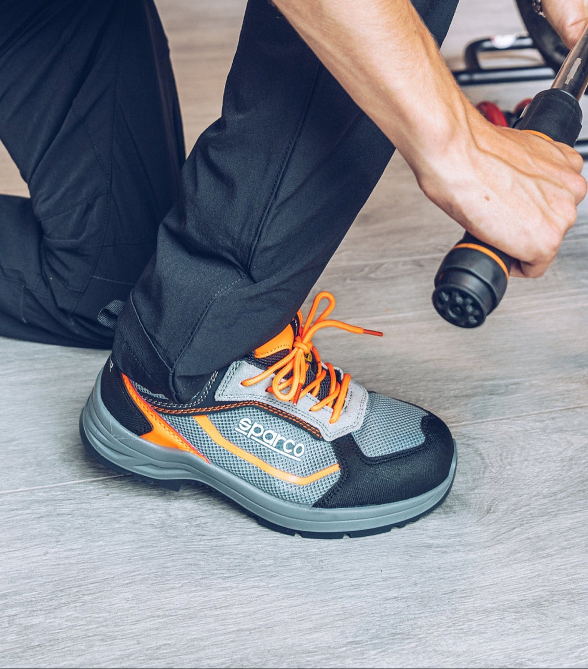 A person kneels on a gray floor wearing a gray and black safety work boot with bright orange laces and orange accents. The person holds a hose nozzle to wet the boot.